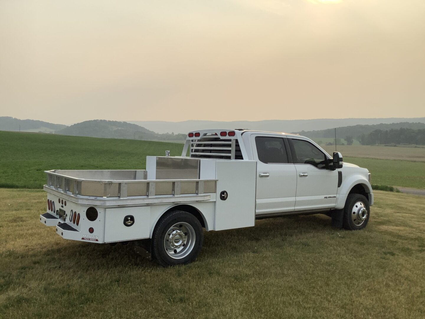 A white pickup truck with a specialized flatbed and storage compartments parked in a field at dusk.