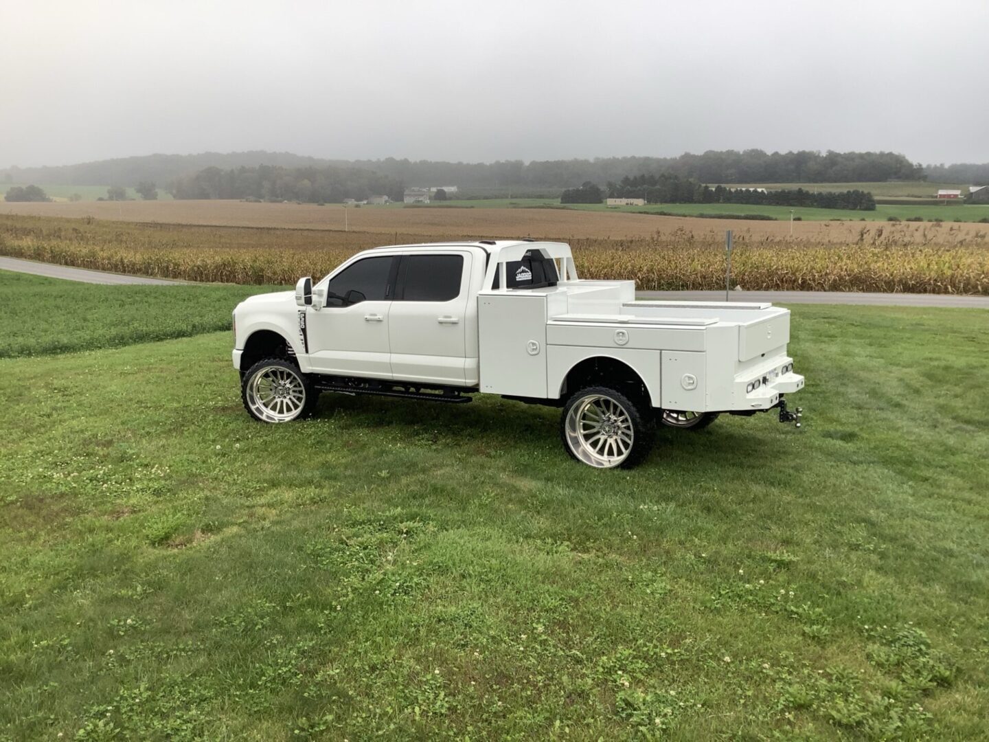 A white pickup truck with custom wheels parked on a grassy knoll near a farm field on an overcast day.
